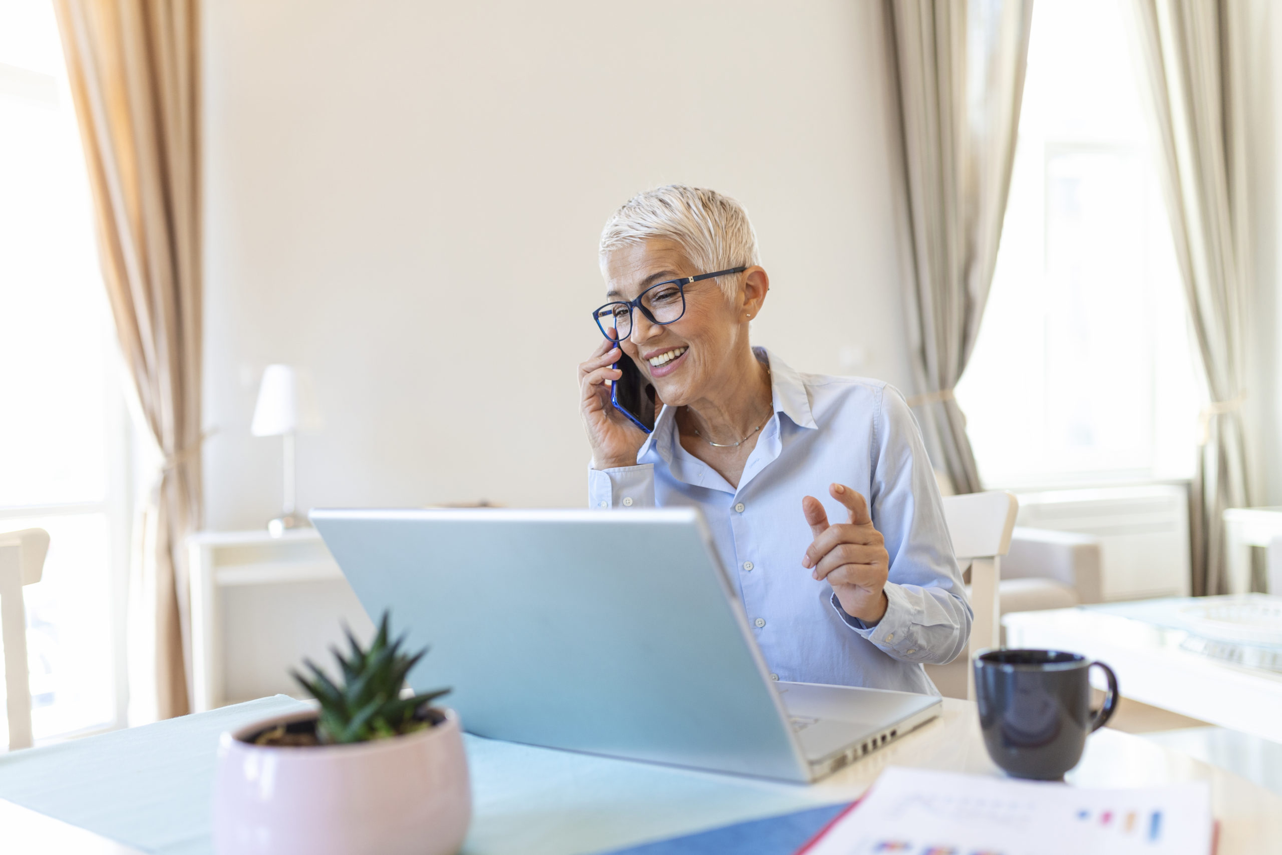 Smiling mature beautiful business woman with white hair working on laptop in bright modern home office.